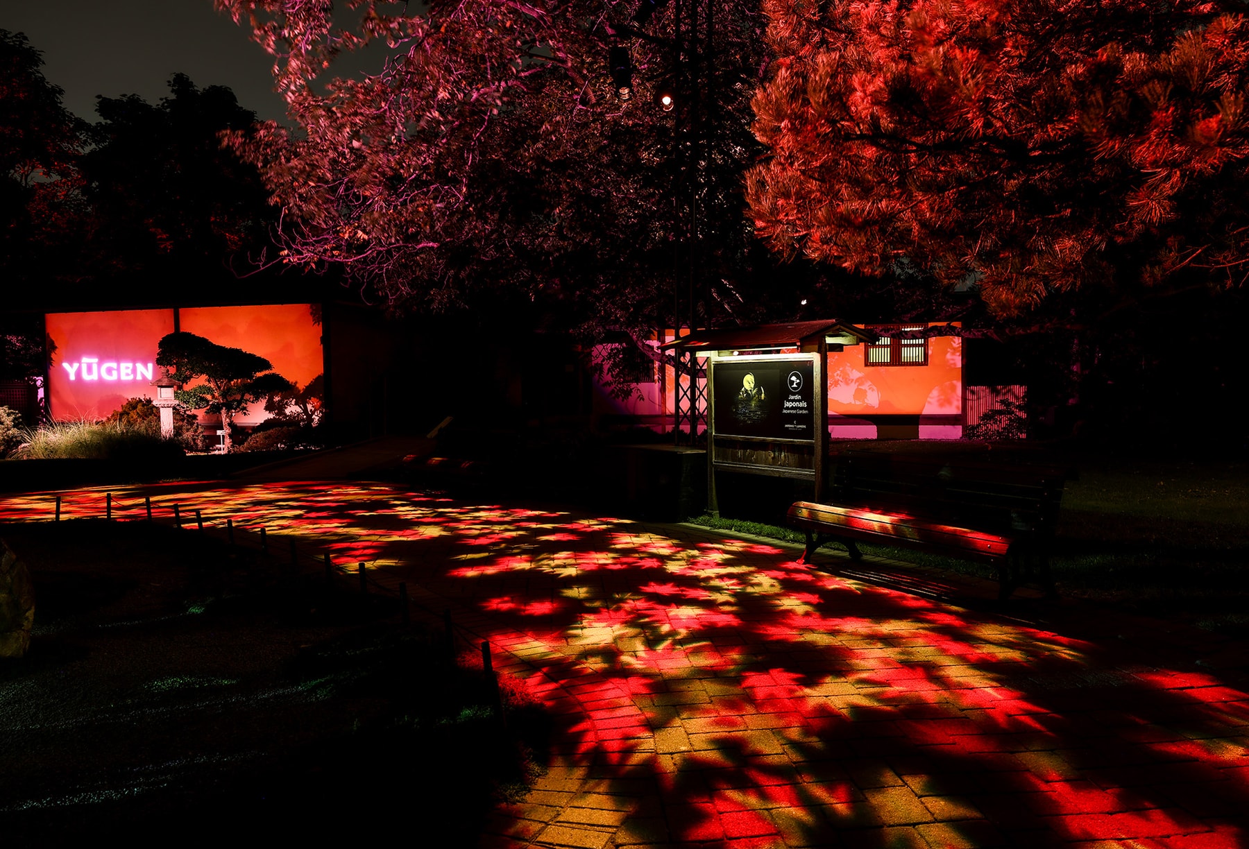 Des motifs lumineux rouges et orange éclairent un sentier près d’un pavillon japonais et d’une enseigne lumineuse « Yūgen ».