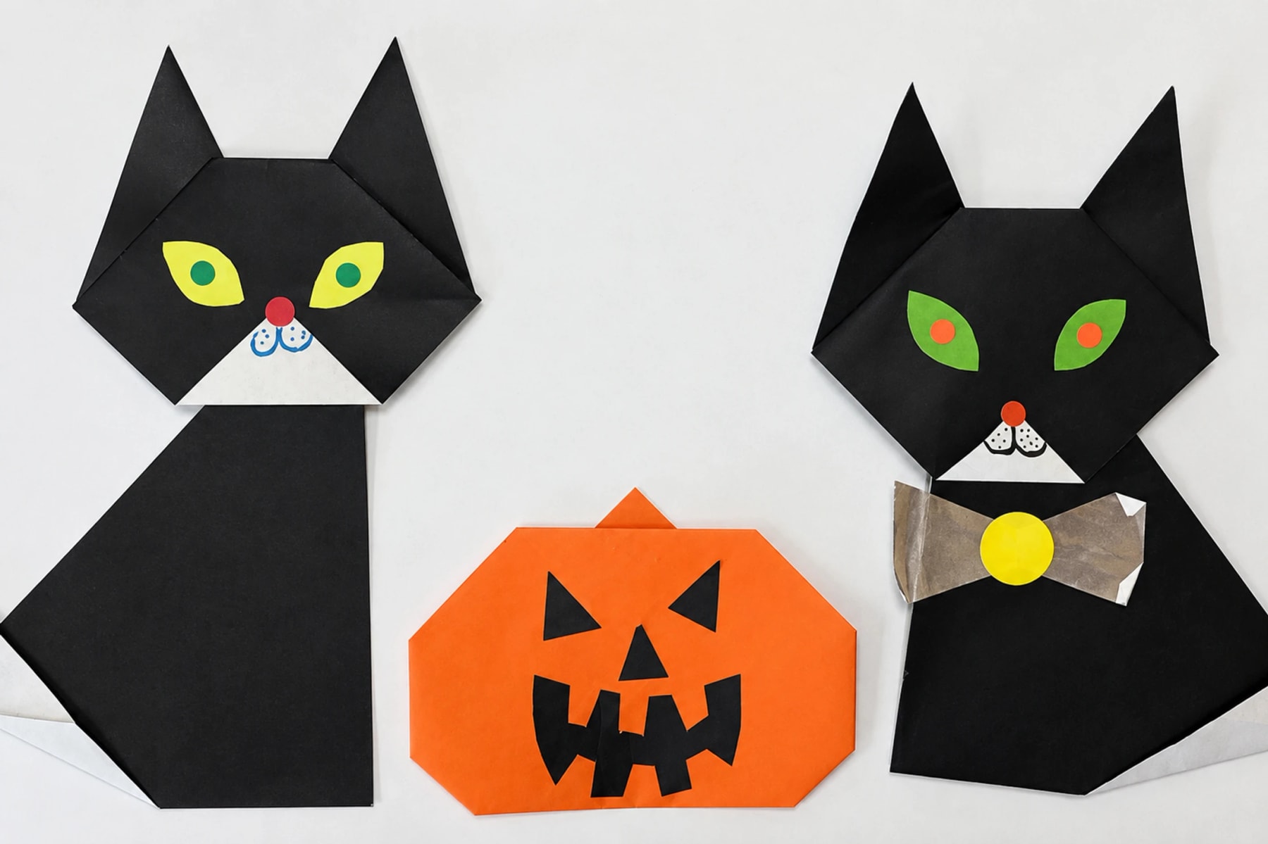 Deux chats noirs en origami à côté d’une citrouille en papier orange.
