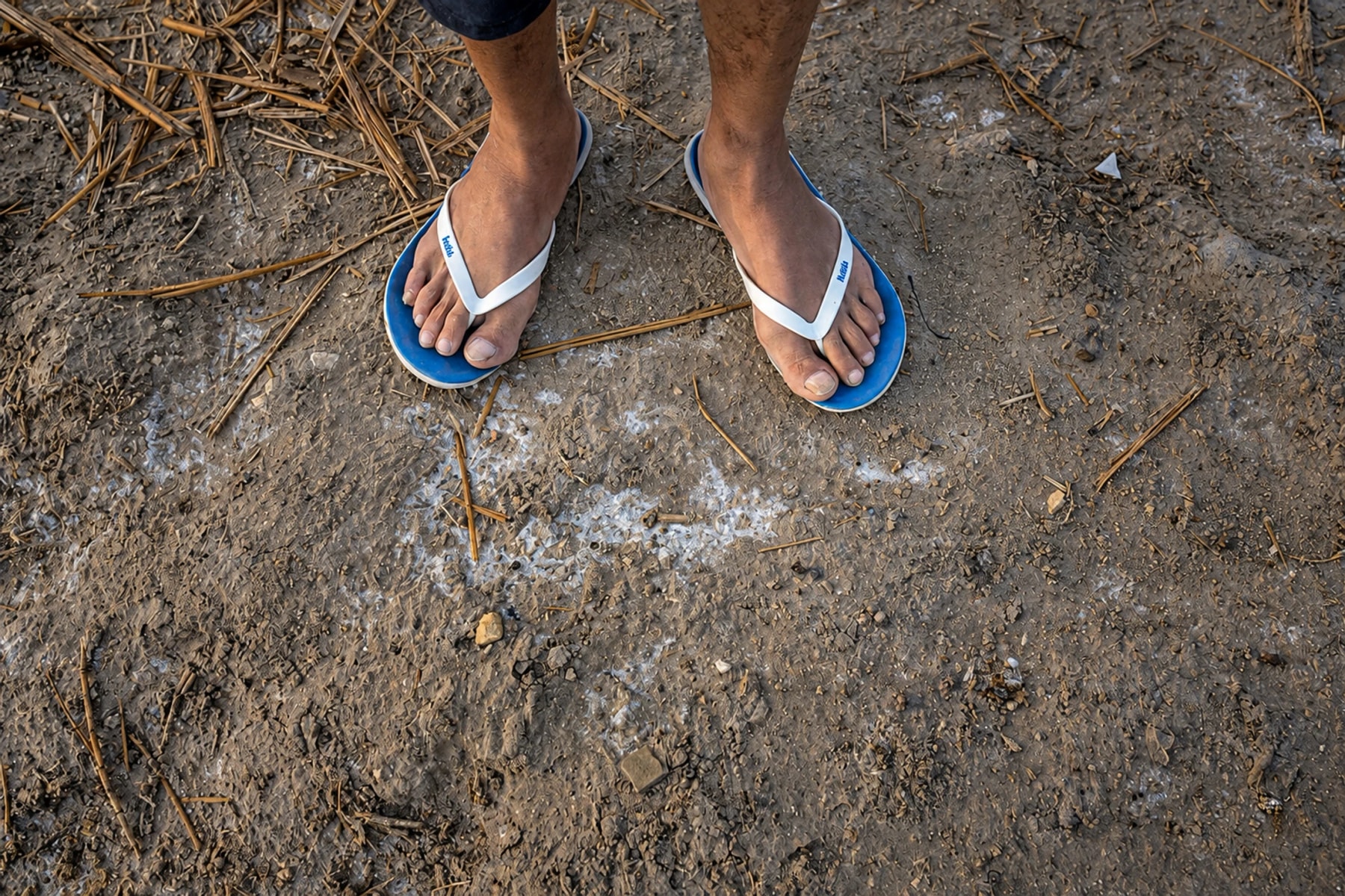 Des pieds chaussés de sandales bleues et blanches sur un sol sec et dénudé.