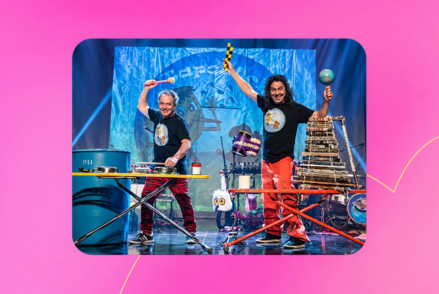 Two musicians hold percussion instruments onstage, framed by a pink border.