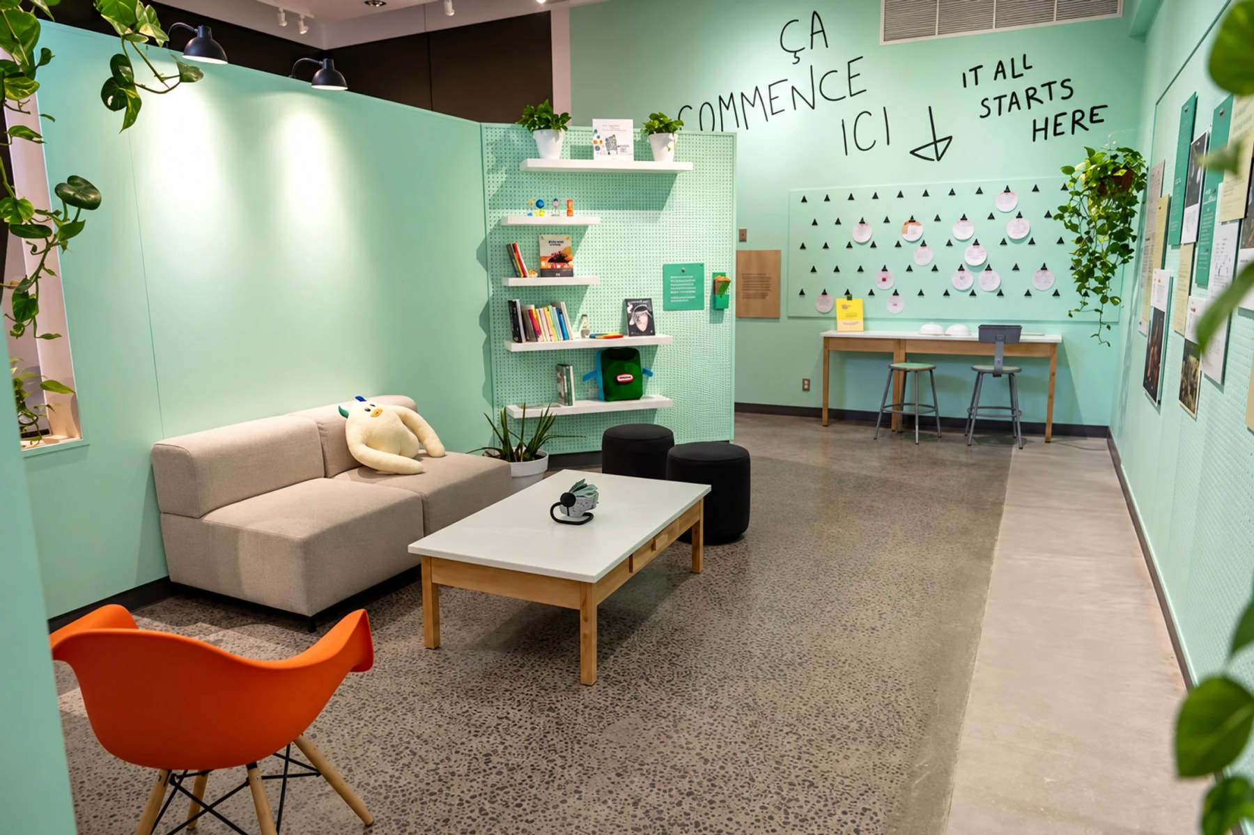 A mint-green exhibition lounge with a sofa, chairs, plants and activity stations.
