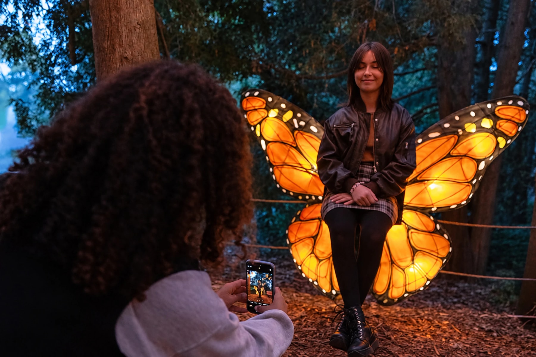 Une personne pose devant des ailes de papillon lumineuses pendant qu’une autre la photographie.