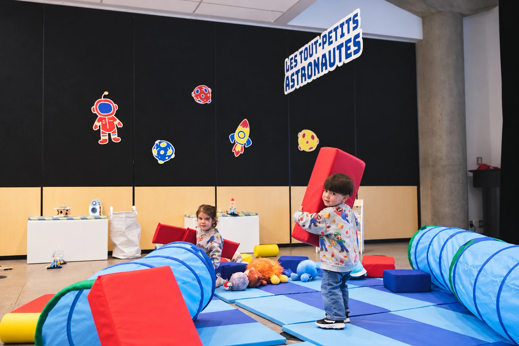 De jeunes enfants jouent avec des blocs de mousse et des tunnels bleus dans une salle sur le thème de l’espace.