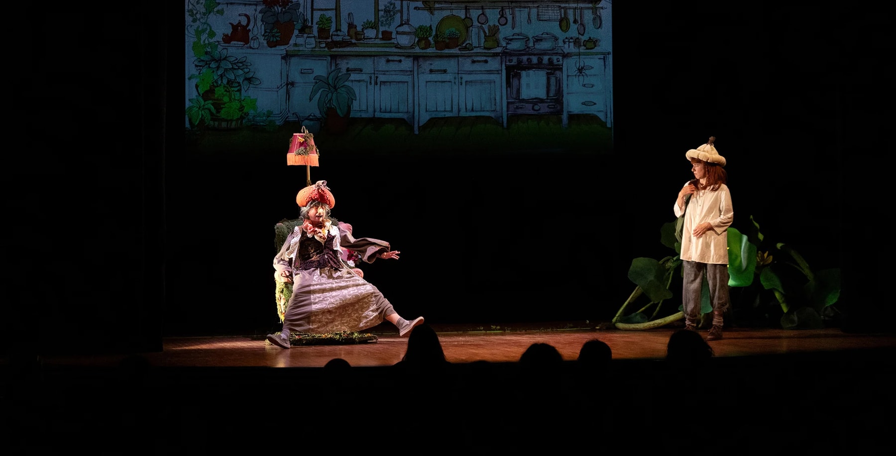 Two costumed performers appear onstage before a projected kitchen scene.
