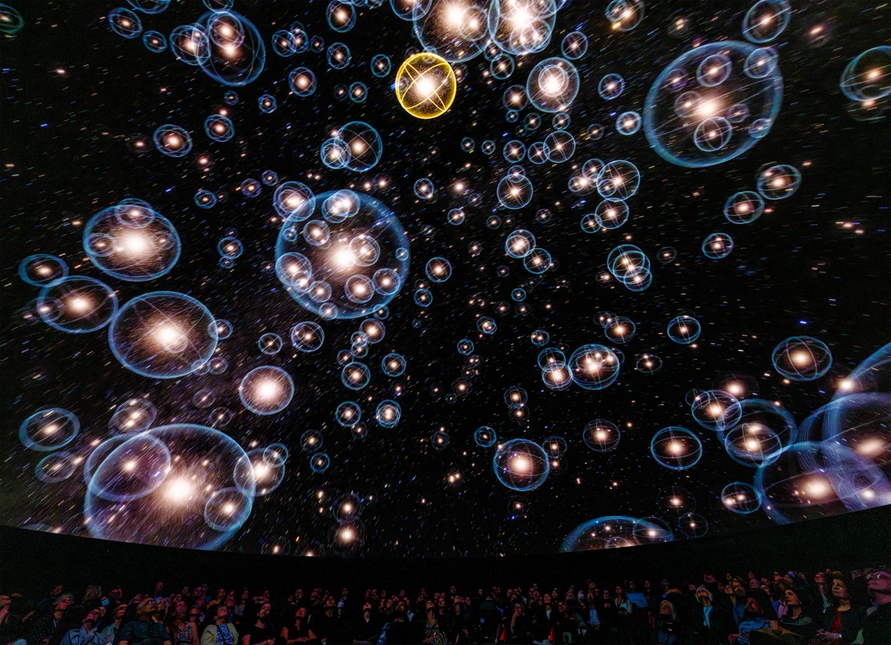 Des sphères lumineuses remplissent le dôme d’un planétarium au-dessus du public assis.