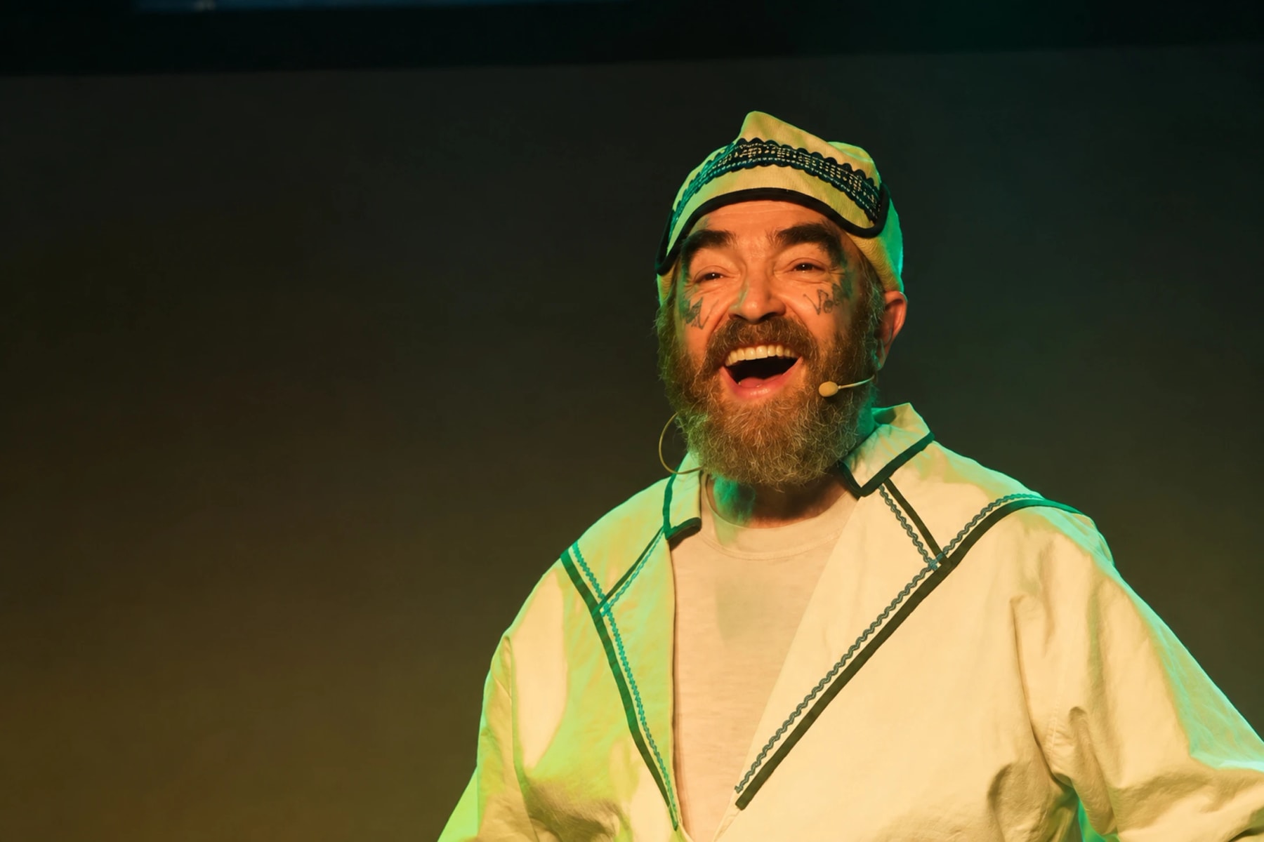 A bearded performer in a decorated tunic speaks onstage.