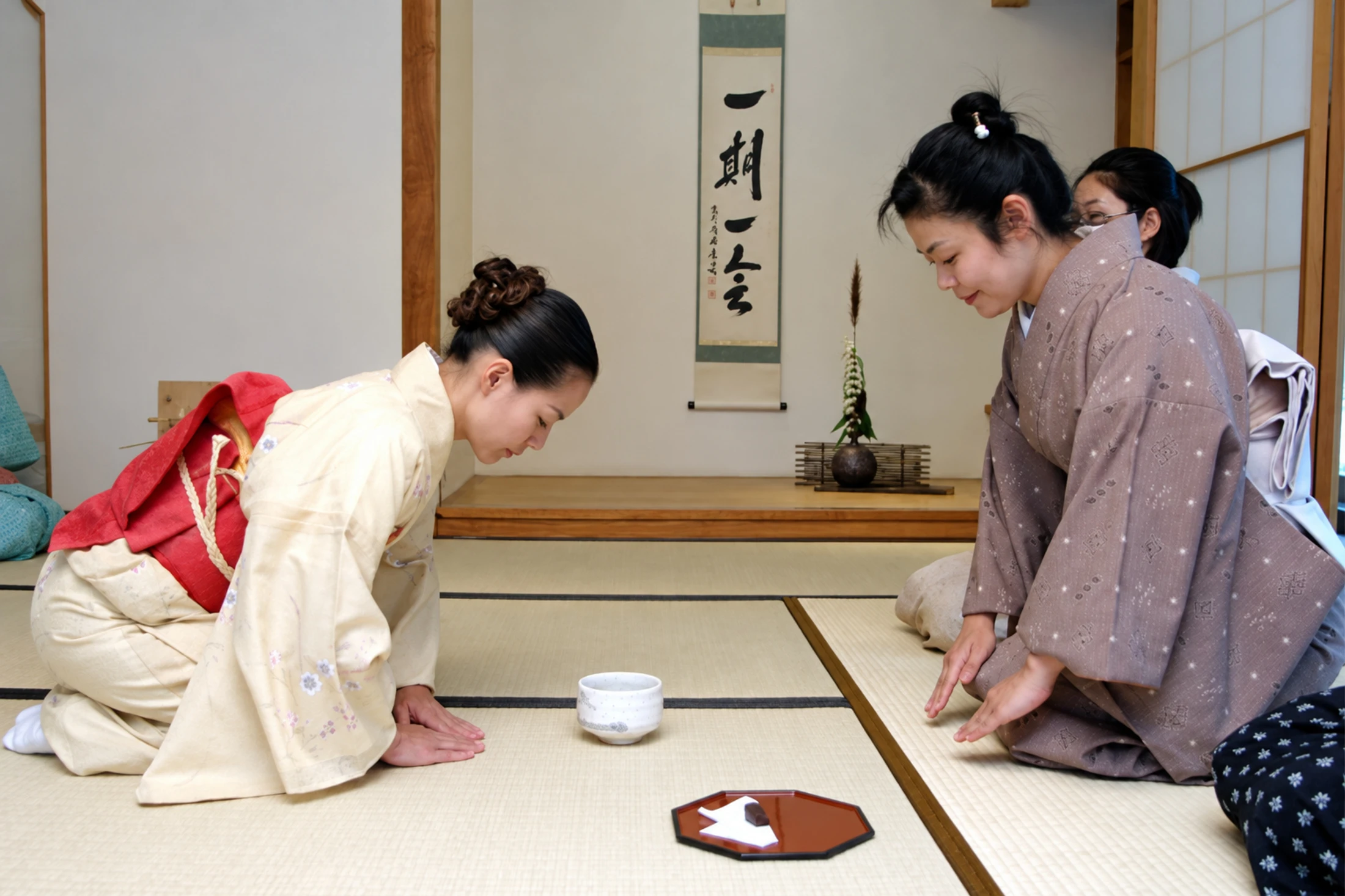 Participants in kimonos bow beside a tea bowl on tatami mats.