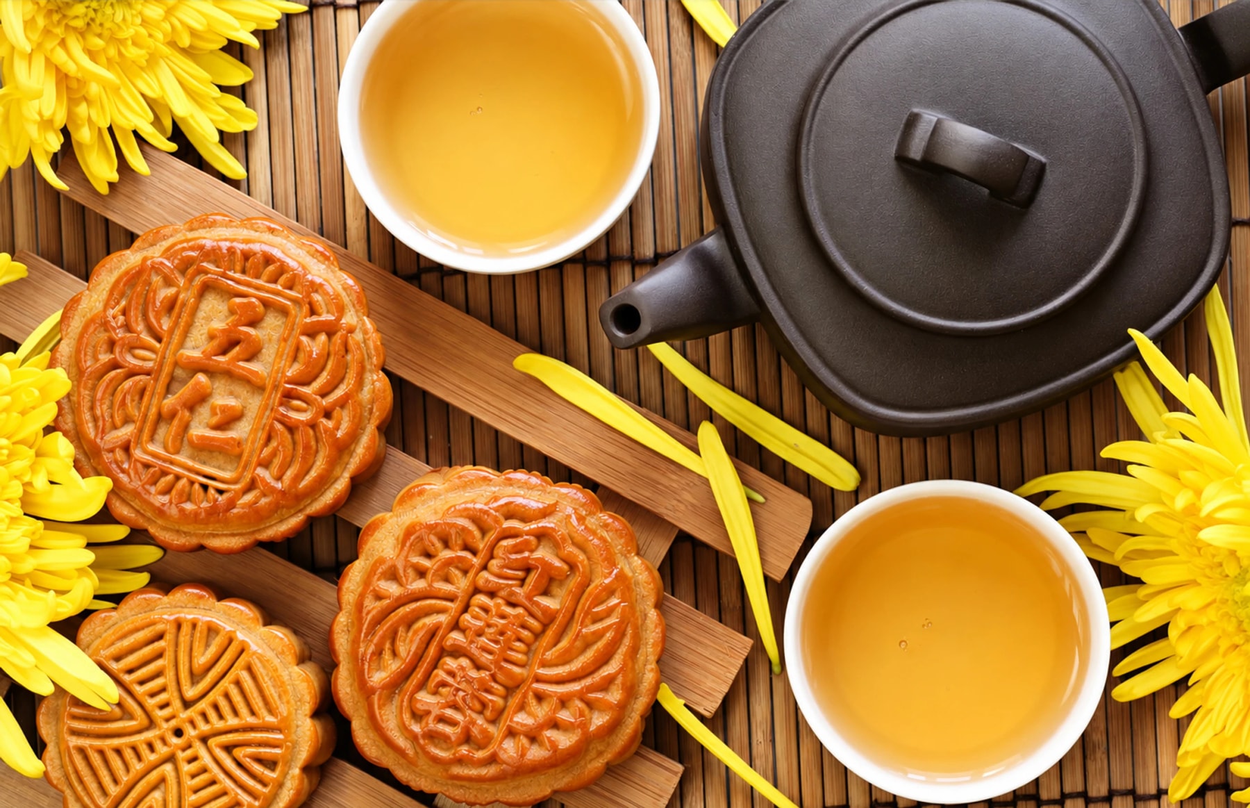 Des gâteaux de lune, des tasses de thé et une théière sombre entourés de fleurs jaunes.