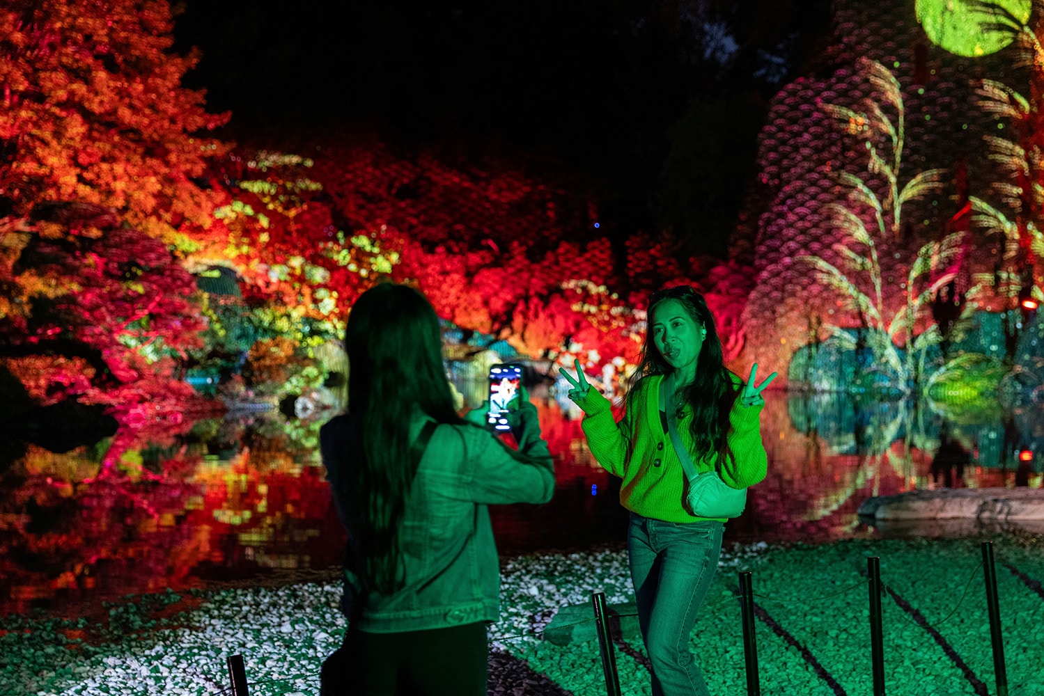Une personne en photographie une autre, qui fait le signe de la paix devant des arbres illuminés la nuit.