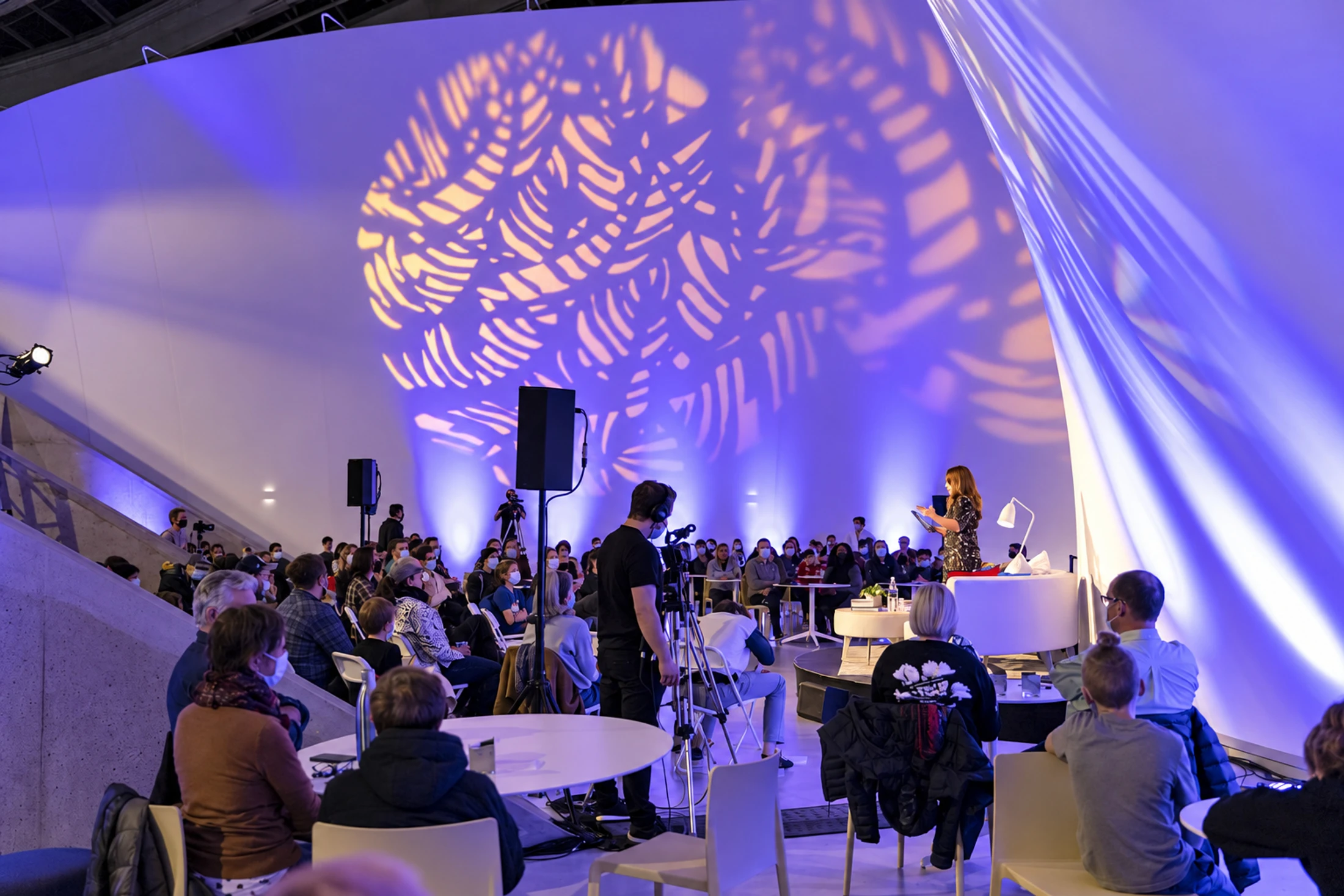 An audience listens to a speaker beneath colourful light projections.