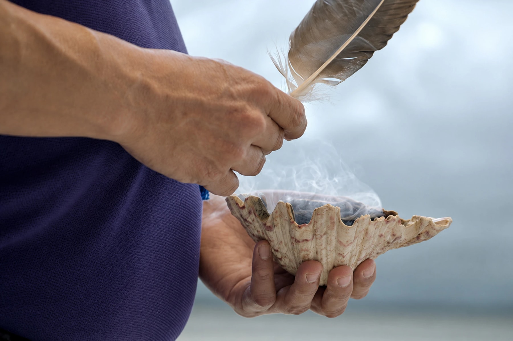 Hands hold a feather above a shell releasing wisps of smoke.
