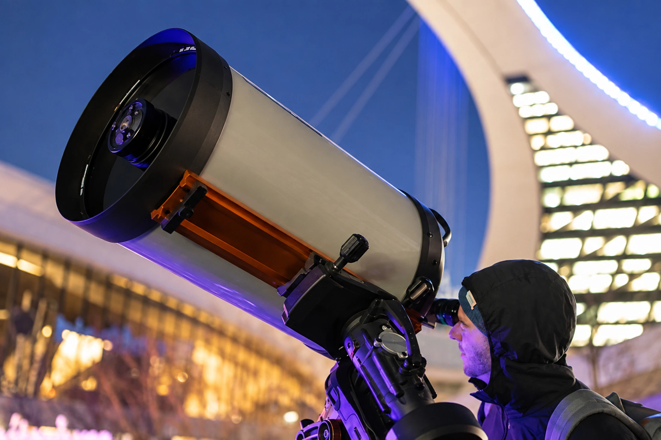 A visitor looks through a telescope with the illuminated Olympic Tower behind.