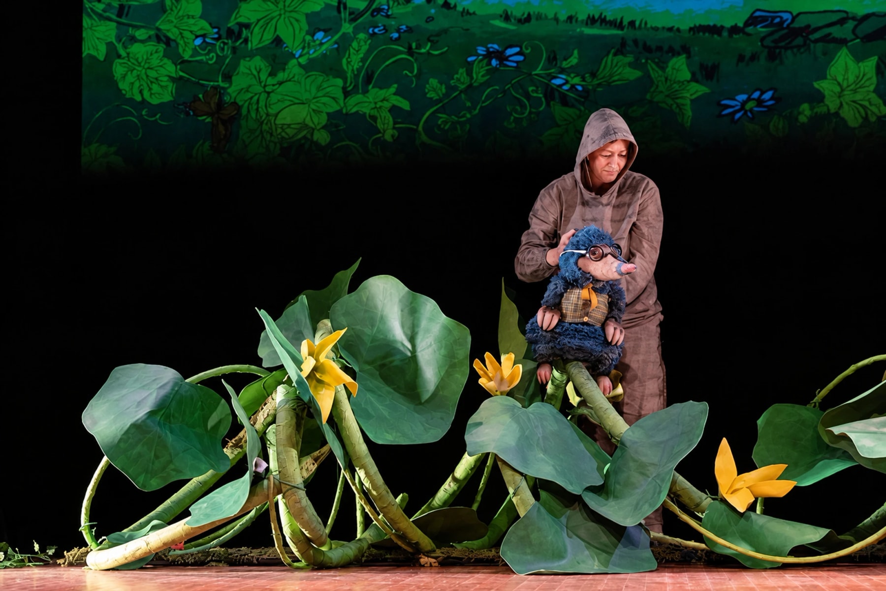 A puppeteer holds a small puppet behind oversized leaves and yellow flowers.