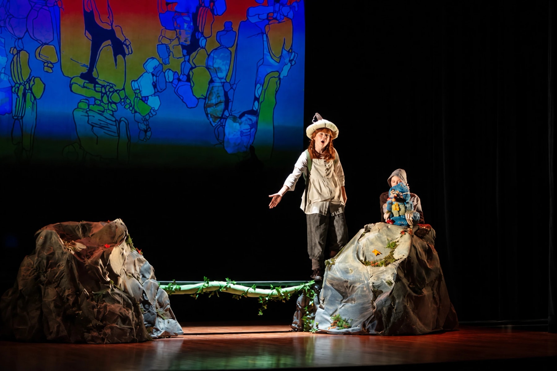 Two performers on a rocky stage set, one holding a puppet, beneath a colourful projection.