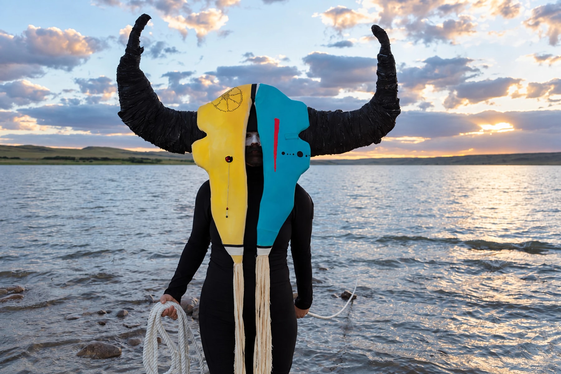 Une personne portant un grand masque à cornes se tient dans l’eau au coucher du soleil.