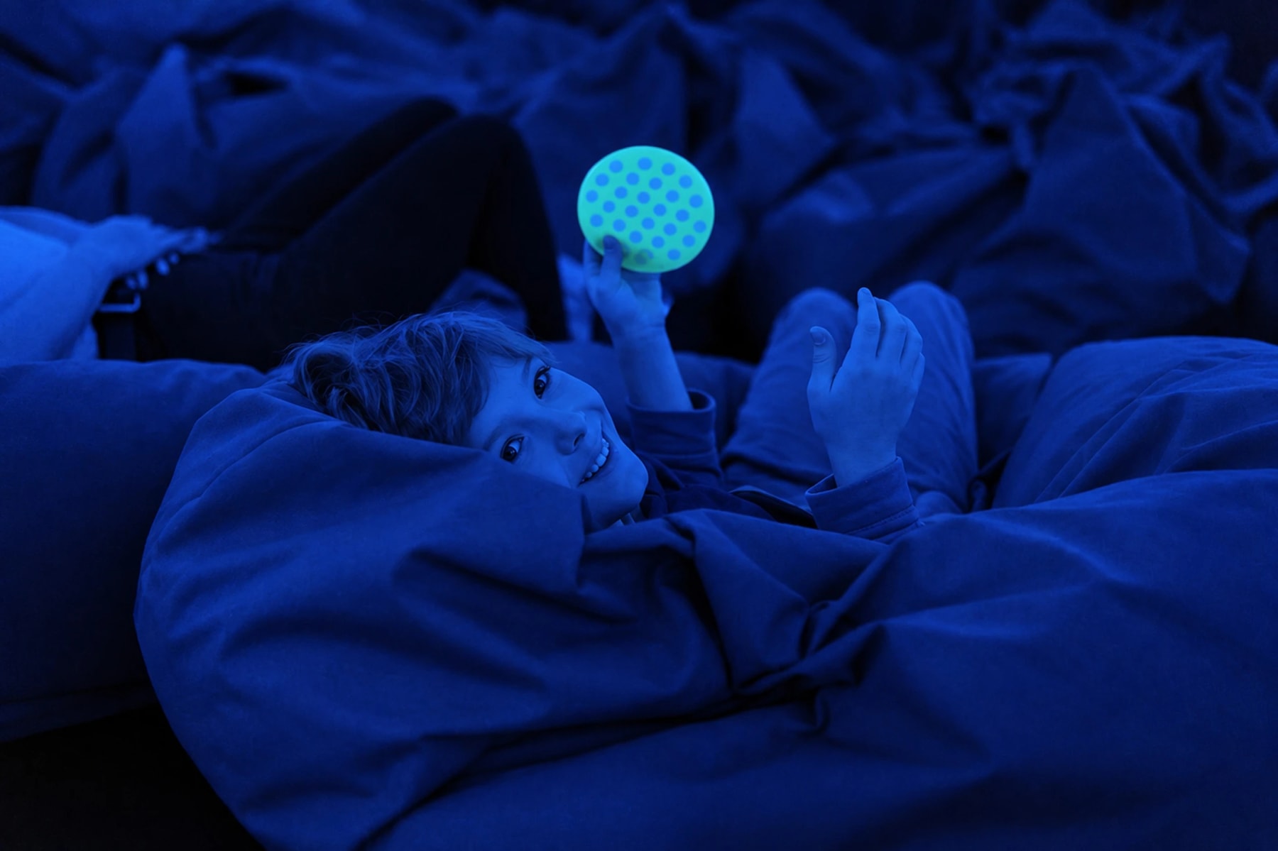 Un enfant allongé sur des coussins tient un disque sensoriel sous une lumière bleue.