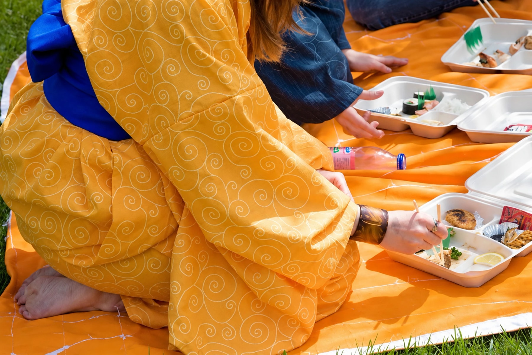 A person in a yellow kimono kneels beside picnic meals on an orange blanket.